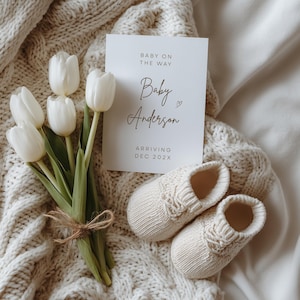 May include: A pregnancy announcement card with the text "Baby on the Way" and "Baby Anderson" is displayed next to a pair of cream-colored baby booties and a bouquet of white tulips. The card also says "Arriving Dec 202X".