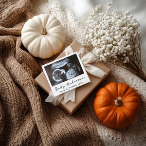 May include: A gift box with a white ribbon, an ultrasound photo that reads "Baby Anderson DUE OCTOBER 202X", two pumpkins, a bouquet of white flowers, and a white pumpkin on a brown knitted blanket.
