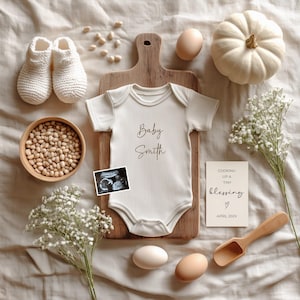 Peut inclure: Une composition à plat avec un body de bébé crème portant l'inscription "Baby Smith", une échographie et une carte indiquant "Cooking up a tiny blessing" avec la date d'avril 202X. L'image comprend également des chaussons de bébé blancs, une petite citrouille, des œufs et un bol en bois.