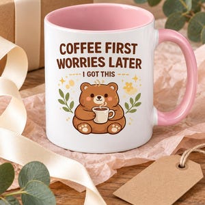 Op de afbeelding: Een witte keramische mok met een roze binnenkant en handvat, voorzien van een illustratie van een bruine beer die een klein kopje vasthoudt. De tekst COFFEE FIRST WORRIES LATER I GOT THIS staat in bruine letters, omringd door sterren en blaadjes.