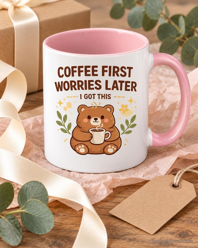 Op de afbeelding: Een witte keramische mok met een roze binnenkant en handvat, voorzien van een illustratie van een bruine beer die een klein kopje vasthoudt. De tekst COFFEE FIRST WORRIES LATER I GOT THIS staat in bruine letters, omringd door sterren en blaadjes.