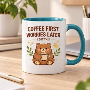 Op de afbeelding: Een witte keramische mok met een blauwgroene binnenkant en handvat, voorzien van een illustratie van een bruine beer met een klein kopje. De tekst COFFEE FIRST WORRIES LATER I GOT THIS staat in bruine letters, omringd door gele sterren en blaadjes.