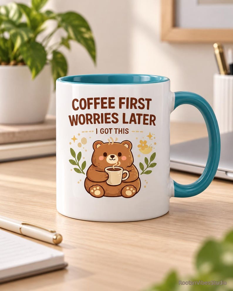Op de afbeelding: Een witte keramische mok met een blauwgroene binnenkant en handvat, voorzien van een illustratie van een bruine beer met een klein kopje. De tekst COFFEE FIRST WORRIES LATER I GOT THIS staat in bruine letters, omringd door gele sterren en blaadjes.