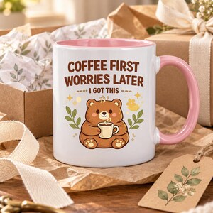 Op de afbeelding: Een witte keramische mok met een roze binnenkant en handvat. De voorzijde toont een illustratie van een bruine beer met een klein kopje, omringd door groene bladeren en gele sterretjes. De tekst luidt: COFFEE FIRST WORRIES LATER I GOT THIS.