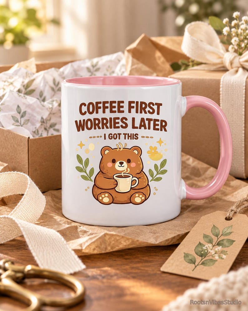Op de afbeelding: Een witte keramische mok met een roze binnenkant en handvat. De voorzijde toont een illustratie van een bruine beer met een klein kopje, omringd door groene bladeren en gele sterretjes. De tekst luidt: COFFEE FIRST WORRIES LATER I GOT THIS.