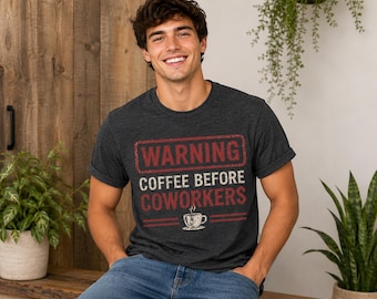 Op de afbeelding: Een donker antracietgrijs t-shirt met korte mouwen en een verweerde rood-witte grafische print met de tekst WARNING COFFEE BEFORE COWORKERS en een kleine illustratie van een dampende koffiekop onder de tekst.