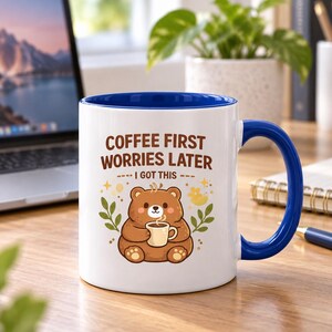 Op de afbeelding: Een witte keramische mok met een blauwe binnenkant en handvat. De voorzijde toont een illustratie van een bruine beer die een klein kopje vasthoudt, omringd door bladeren en sterren. De tekst luidt: COFFEE FIRST WORRIES LATER I GOT THIS in bruin.