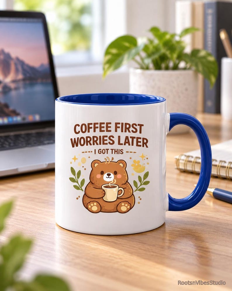 Op de afbeelding: Een witte keramische mok met een blauwe binnenkant en handvat. De voorzijde toont een illustratie van een bruine beer die een klein kopje vasthoudt, omringd door bladeren en sterren. De tekst luidt: COFFEE FIRST WORRIES LATER I GOT THIS in bruin.