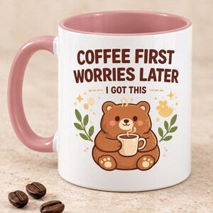 Op de afbeelding: Een witte keramische mok met een oudroze binnenkant en handvat. De voorzijde toont een illustratie van een bruine beer die een klein kopje vasthoudt, omringd door blaadjes en sterretjes. De tekst luidt: COFFEE FIRST WORRIES LATER I GOT THIS in bruin.