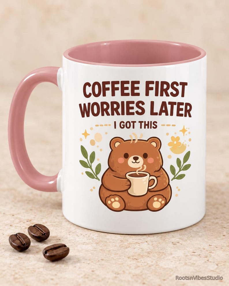 Op de afbeelding: Een witte keramische mok met een oudroze binnenkant en handvat. De voorzijde toont een illustratie van een bruine beer die een klein kopje vasthoudt, omringd door blaadjes en sterretjes. De tekst luidt: COFFEE FIRST WORRIES LATER I GOT THIS in bruin.