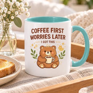 Op de afbeelding: Een witte keramische mok met een lichtblauwe binnenkant en handvat. De voorzijde toont een illustratie van een bruine beer die een klein kopje vasthoudt, met de tekst "COFFEE FIRST WORRIES LATER" en "I GOT THIS" in bruin erboven, omringd door blaadjes.