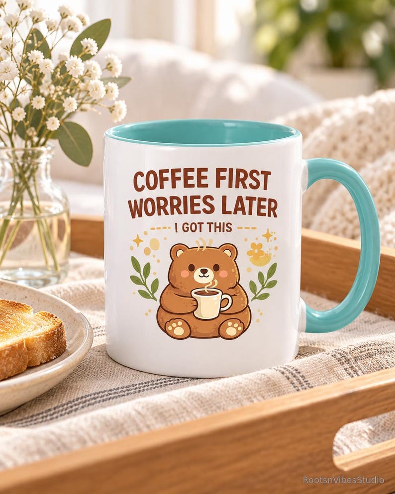 Op de afbeelding: Een witte keramische mok met een lichtblauwe binnenkant en handvat. De voorzijde toont een illustratie van een bruine beer die een klein kopje vasthoudt, met de tekst "COFFEE FIRST WORRIES LATER" en "I GOT THIS" in bruin erboven, omringd door blaadjes.