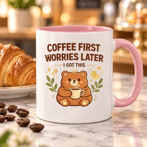 Op de afbeelding: Een witte keramische mok met een roze binnenkant en handvat, voorzien van een illustratie van een bruine beer die een klein kopje vasthoudt. De tekst luidt COFFEE FIRST WORRIES LATER I GOT THIS in bruine letters. Het ontwerp bevat kleine groene blaadjes en gele sterren.