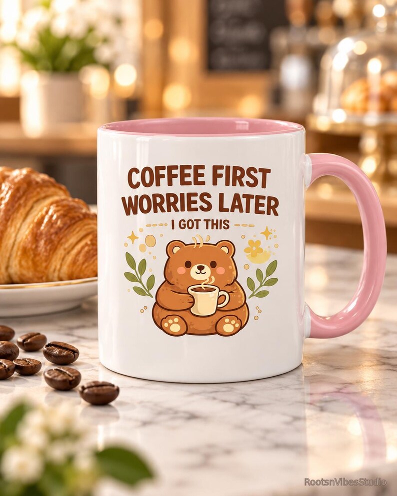 Op de afbeelding: Een witte keramische mok met een roze binnenkant en handvat, voorzien van een illustratie van een bruine beer die een klein kopje vasthoudt. De tekst luidt COFFEE FIRST WORRIES LATER I GOT THIS in bruine letters. Het ontwerp bevat kleine groene blaadjes en gele sterren.