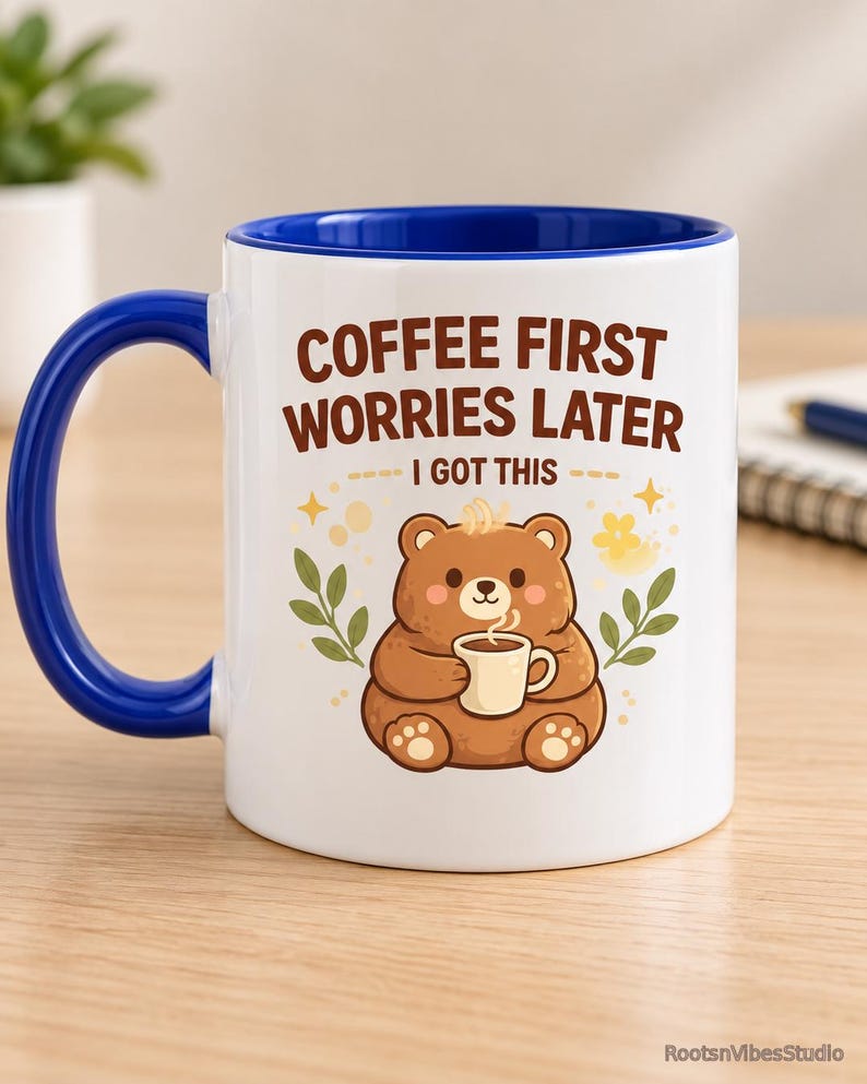 Op de afbeelding: Een witte keramische mok met een blauw handvat en binnenrand. De voorzijde toont een illustratie van een bruine beer die een klein kopje vasthoudt, omringd door groene bladeren en gele sterren. De tekst luidt: COFFEE FIRST WORRIES LATER I GOT THIS.