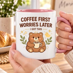 Op de afbeelding: Een witte keramische mok met een roze binnenkant en handvat, voorzien van een illustratie van een bruine beer die een klein kopje koffie vasthoudt. De tekst luidt COFFEE FIRST WORRIES LATER en I GOT THIS, omringd door gele sterretjes en groene takjes.