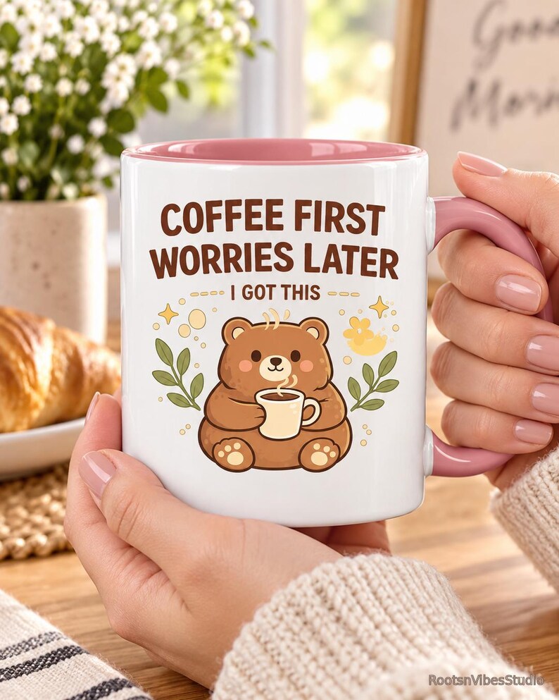 Op de afbeelding: Een witte keramische mok met een roze binnenkant en handvat, voorzien van een illustratie van een bruine beer die een klein kopje koffie vasthoudt. De tekst luidt COFFEE FIRST WORRIES LATER en I GOT THIS, omringd door gele sterretjes en groene takjes.