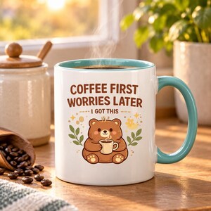 Op de afbeelding: Een witte keramische mok met een talingkleurige binnenkant en handvat, voorzien van een bruine beer die een klein kopje vasthoudt. De tekst luidt COFFEE FIRST WORRIES LATER I GOT THIS. Stoom stijgt op uit de mok met een donkere drank.