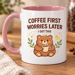 Op de afbeelding: Een witte keramische mok met een roze handvat en binnenkant. De voorzijde toont een illustratie van een bruine beer die een klein kopje vasthoudt, omringd door blaadjes en sterren. De tekst luidt: COFFEE FIRST WORRIES LATER I GOT THIS in bruin.