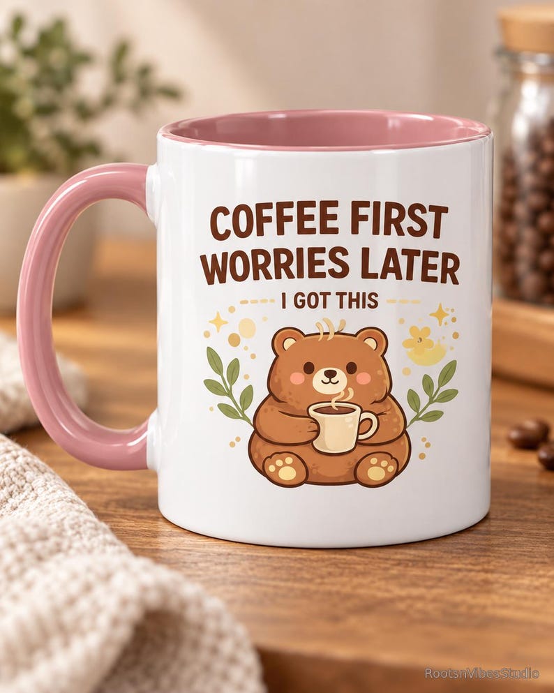 Op de afbeelding: Een witte keramische mok met een roze handvat en binnenkant. De voorzijde toont een illustratie van een bruine beer die een klein kopje vasthoudt, omringd door blaadjes en sterren. De tekst luidt: COFFEE FIRST WORRIES LATER I GOT THIS in bruin.
