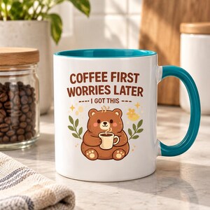 Op de afbeelding: Een witte keramische mok met een blauwgroene binnenkant en handvat, voorzien van een illustratie van een bruine beer met een klein kopje, en de tekst "COFFEE FIRST WORRIES LATER" en "I GOT THIS" in bruin, omringd door kleine blaadjes en sterren.