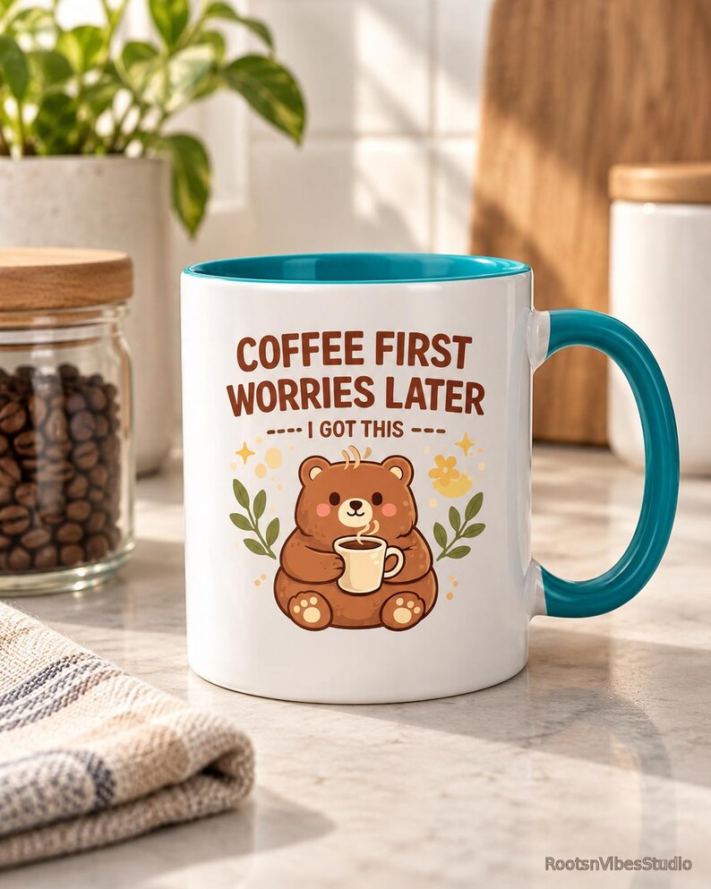 Op de afbeelding: Een witte keramische mok met een blauwgroene binnenkant en handvat, voorzien van een illustratie van een bruine beer met een klein kopje, en de tekst "COFFEE FIRST WORRIES LATER" en "I GOT THIS" in bruin, omringd door kleine blaadjes en sterren.