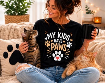 Op de afbeelding: Een zwart t-shirt met de witte en oranje tekst "MY KIDS HAVE PAWS", grafische pootafdrukken en een klein rood hartje. Het shirt wordt gedragen met een lichtblauwe spijkerbroek en het ontwerp bevat decoratieve lijnen en kleine hartjes.