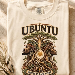 May include: A cream-colored t-shirt featuring a graphic print with the text UBUNTU and ROOTED IN EACH OTHER. The design depicts two profiles facing outward, a large tree with exposed roots, and a central sun icon in shades of green, brown, and gold.