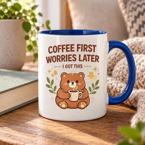 Op de afbeelding: Een witte keramische mok met een blauwe binnenkant en handvat, voorzien van een illustratie van een bruine beer die een klein kopje koffie vasthoudt. De tekst luidt COFFEE FIRST WORRIES LATER I GOT THIS, met gele sterren en bladeren.