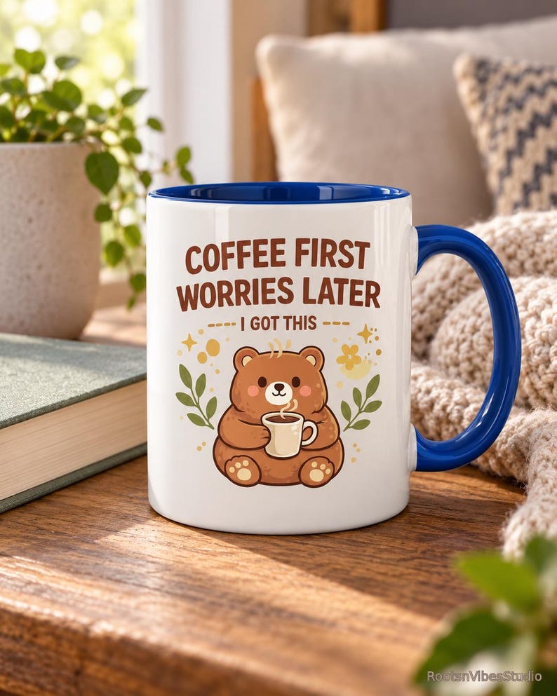 Op de afbeelding: Een witte keramische mok met een blauwe binnenkant en handvat, voorzien van een illustratie van een bruine beer die een klein kopje koffie vasthoudt. De tekst luidt COFFEE FIRST WORRIES LATER I GOT THIS, met gele sterren en bladeren.