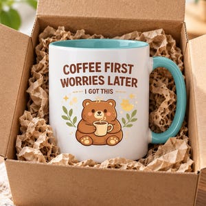 Op de afbeelding: Een witte keramische mok met een lichtblauwe binnenkant en handvat, voorzien van een bruine beer die een klein kopje vasthoudt. De tekst COFFEE FIRST WORRIES LATER I GOT THIS staat in bruine letters, omringd door gele sterren en groene blaadjes.