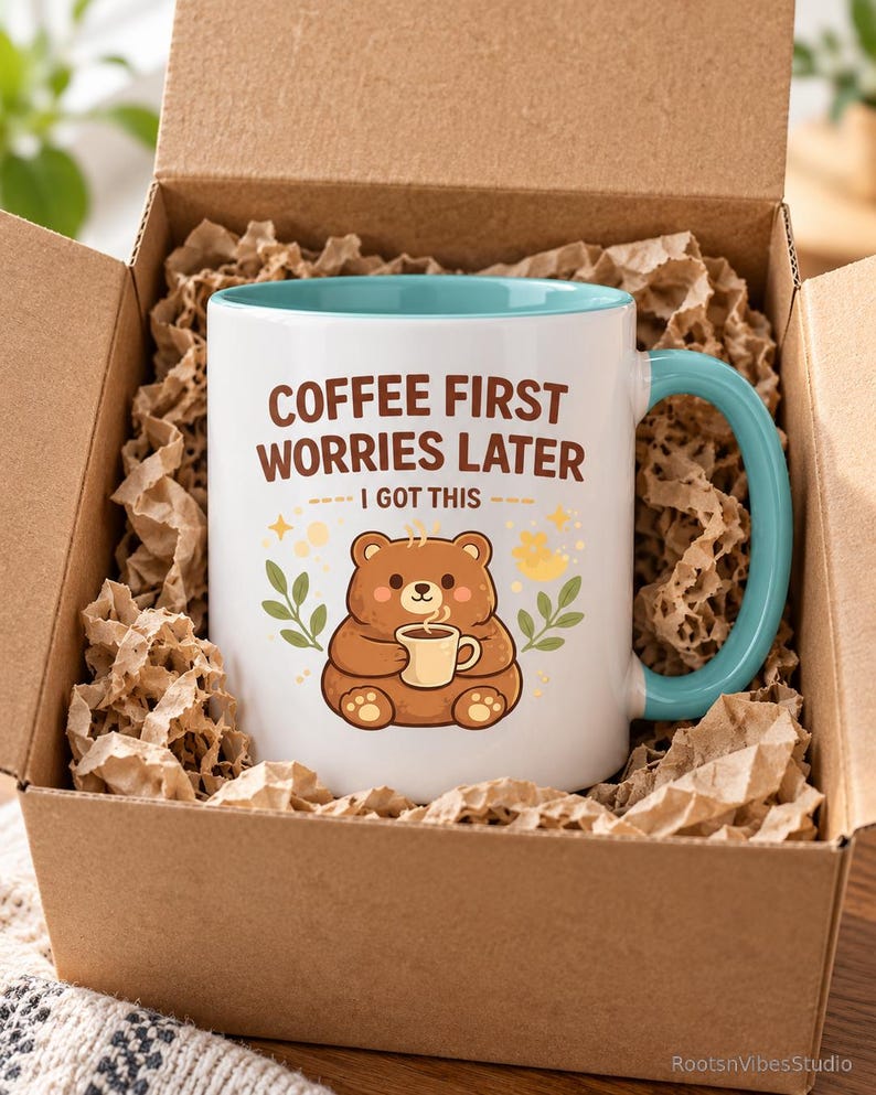 Op de afbeelding: Een witte keramische mok met een lichtblauwe binnenkant en handvat, voorzien van een bruine beer die een klein kopje vasthoudt. De tekst COFFEE FIRST WORRIES LATER I GOT THIS staat in bruine letters, omringd door gele sterren en groene blaadjes.