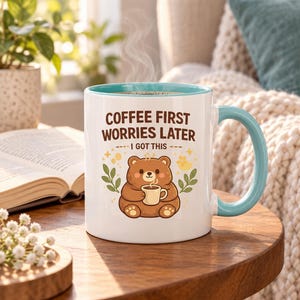 Op de afbeelding: Een witte keramische mok met een lichtblauwe binnenkant en handvat, voorzien van een illustratie van een bruine beer die een klein kopje vasthoudt, omringd door groene bladeren en gele accenten, met de tekst COFFEE FIRST WORRIES LATER I GOT THIS.