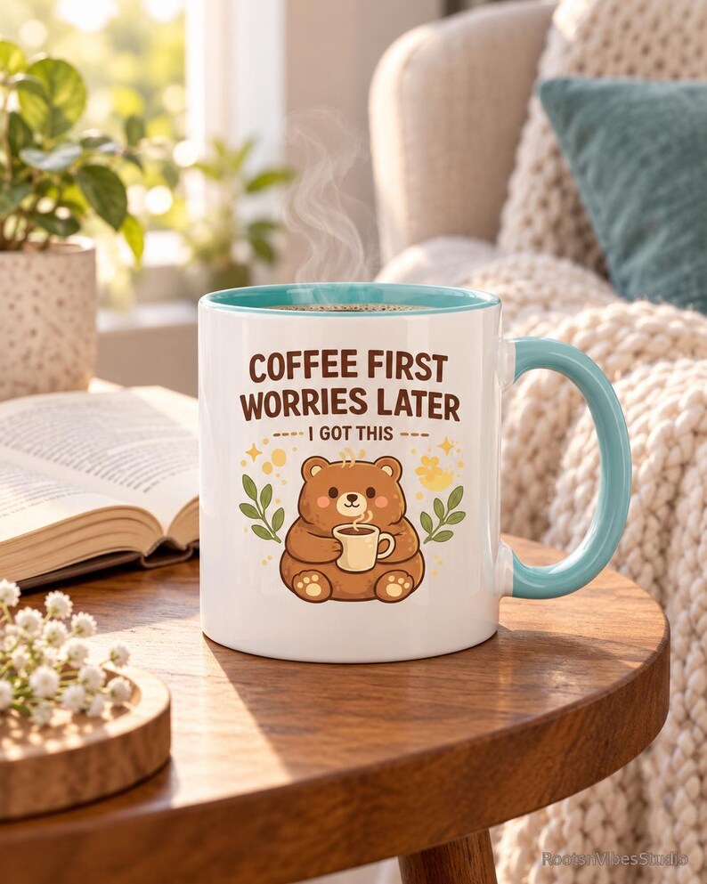 Op de afbeelding: Een witte keramische mok met een lichtblauwe binnenkant en handvat, voorzien van een illustratie van een bruine beer die een klein kopje vasthoudt, omringd door groene bladeren en gele accenten, met de tekst COFFEE FIRST WORRIES LATER I GOT THIS.