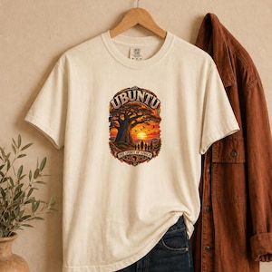 May include: An off-white short-sleeve t-shirt featuring a graphic print of a sunset landscape with a large tree, birds, and silhouettes of people. The text reads "UBUNTU" at the top and "THE SPIRIT OF AFRICA" at the bottom of the oval design.