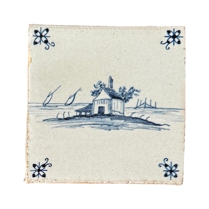 May include: Square, off-white ceramic tile with a hand-painted blue and white landscape scene. The scene depicts a small building on an island, with stylized trees and water. Decorative floral corner accents complete the design.