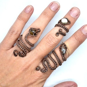 May include: Three copper wire rings with gemstone accents. The rings are shaped like snakes with the gemstones set in the head of each snake. The gemstones are a brown tiger's eye, a white pearl, and a clear crystal.