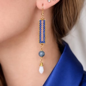 May include: Gold-tone earrings with a rectangular blue beaded design. The earrings have a gold frame with a row of blue beads inside. Below the rectangle is a blue and gray bead, and a white teardrop-shaped bead.