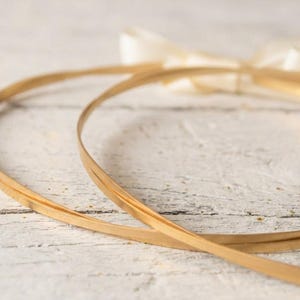 May include: Two thin, gold-colored hoops, possibly for a wreath or craft project, are arranged on a white, weathered wooden surface. A cream-colored ribbon is tied in a bow in the background. The hoops are made of a thin, flexible material.