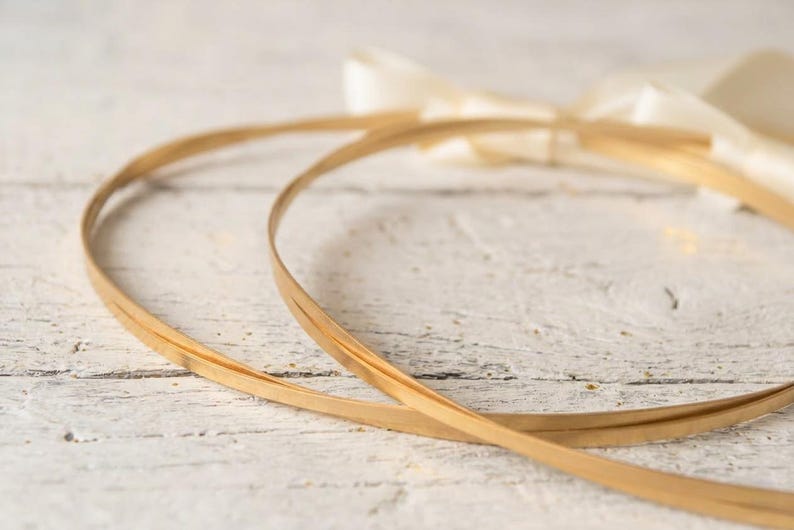 May include: Two thin, gold-colored hoops, possibly for a wreath or craft project, are arranged on a white, weathered wooden surface. A cream-colored ribbon is tied in a bow in the background. The hoops are made of a thin, flexible material.