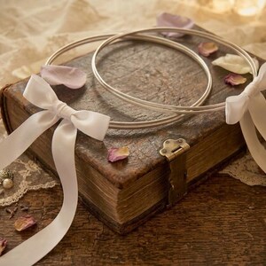May include: Two silver wedding rings rest on a vintage book with a brass clasp. White satin ribbons are tied in bows and draped over the book, with scattered rose petals. The scene is set on a wooden surface.
