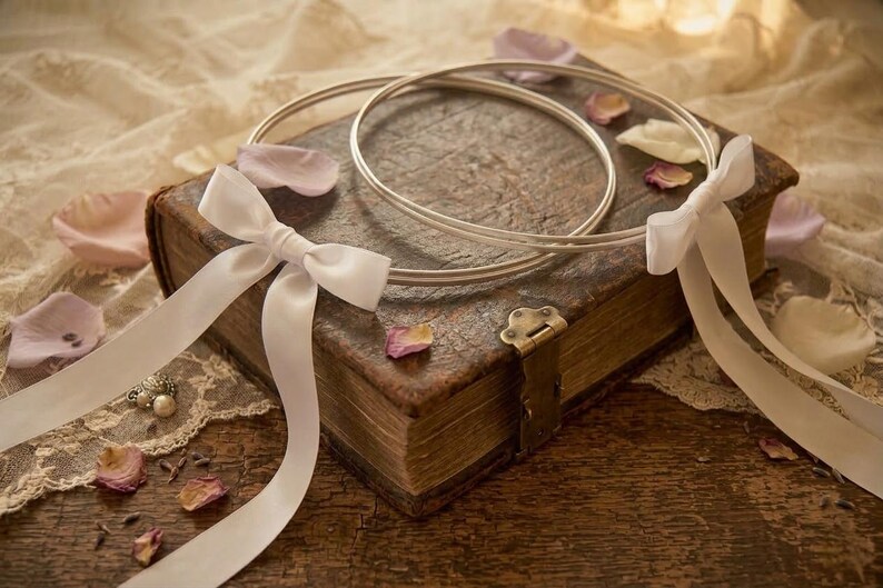 May include: Two silver wedding rings rest on a vintage book with a brass clasp. White satin ribbons are tied in bows and draped over the book, with scattered rose petals. The scene is set on a wooden surface.