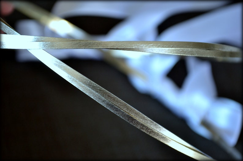 May include: Close-up of silver ribbon, with a metallic sheen, overlapping against a blurred white background. The ribbon appears to be thin and delicate, with a slight texture. The image is well-lit, highlighting the ribbon's reflective surface.