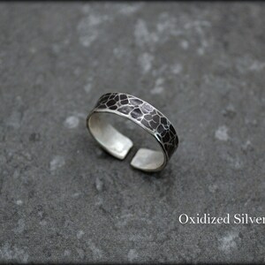 May include: An oxidized silver ring with a textured, geometric pattern. The adjustable ring has a dark, almost black, design on the band. The ring is displayed on a gray surface. The text "Oxidized Silver" is visible.