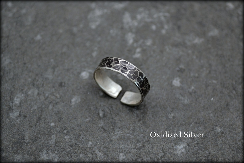 May include: An oxidized silver ring with a textured, geometric pattern. The adjustable ring has a dark, almost black, design on the band. The ring is displayed on a gray surface. The text "Oxidized Silver" is visible.