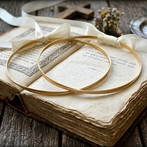 May include: Two gold wedding bands rest on an open antique book with aged pages. A white ribbon and a small bouquet of dried flowers are also present. The scene is set on a rustic wooden surface, creating a vintage aesthetic.