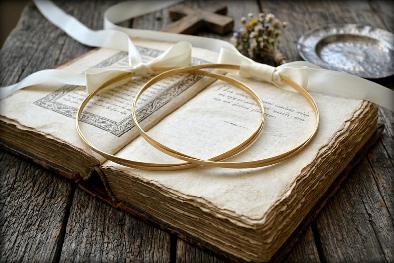May include: Two gold wedding bands rest on an open antique book with aged pages. A white ribbon and a small bouquet of dried flowers are also present. The scene is set on a rustic wooden surface, creating a vintage aesthetic.