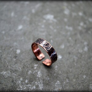 May include: Copper adjustable ring with a hammered texture. The ring features a dark, mottled pattern on the band, creating a unique visual effect. The open design allows for size adjustment. The ring is set against a gray background.