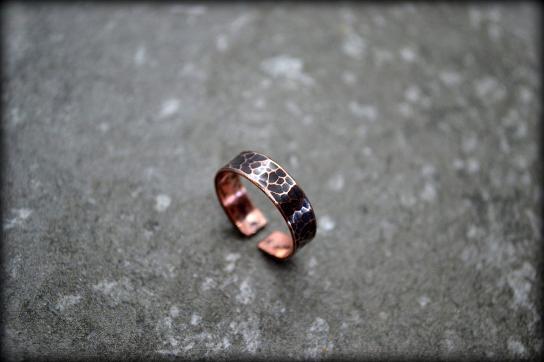 May include: Copper adjustable ring with a hammered texture. The ring features a dark, mottled pattern on the band, creating a unique visual effect. The open design allows for size adjustment. The ring is set against a gray background.