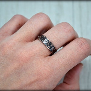 May include: A hammered copper ring with a dark, textured pattern. The ring is worn on a finger, showcasing its unique, handcrafted design. The band has a width of approximately 0.25 inches.